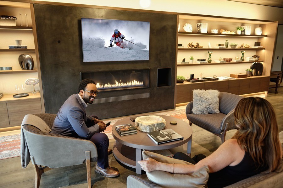 Two people sitting in a living room watching a ski show. A Josh.ai microphone is on the wall next to the TV.