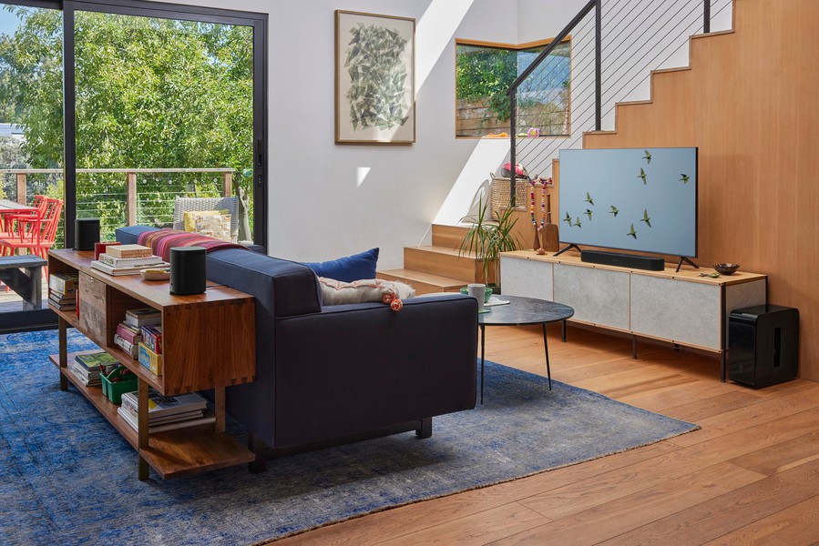 A bright, modern living room features a navy sofa, wood shelving, and a flat-screen TV on a media console, with a Sonos speaker system and sliding glass doors opening to a sunny patio.